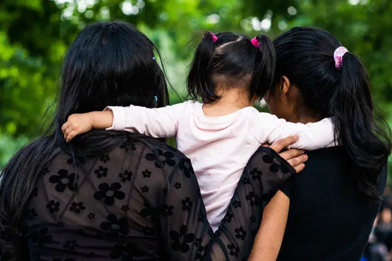 Image of three people's backs, two of them hug a kid who's in the middle at Maria Hernandez Park in Brooklyn