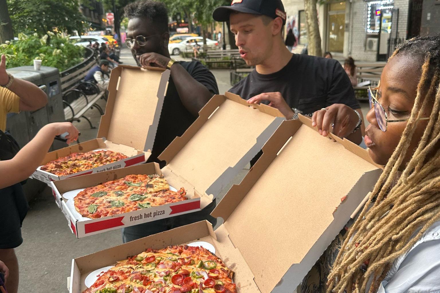 Three people stand on a sidewalk, with pizza boxes open, revealing pizzas inside.