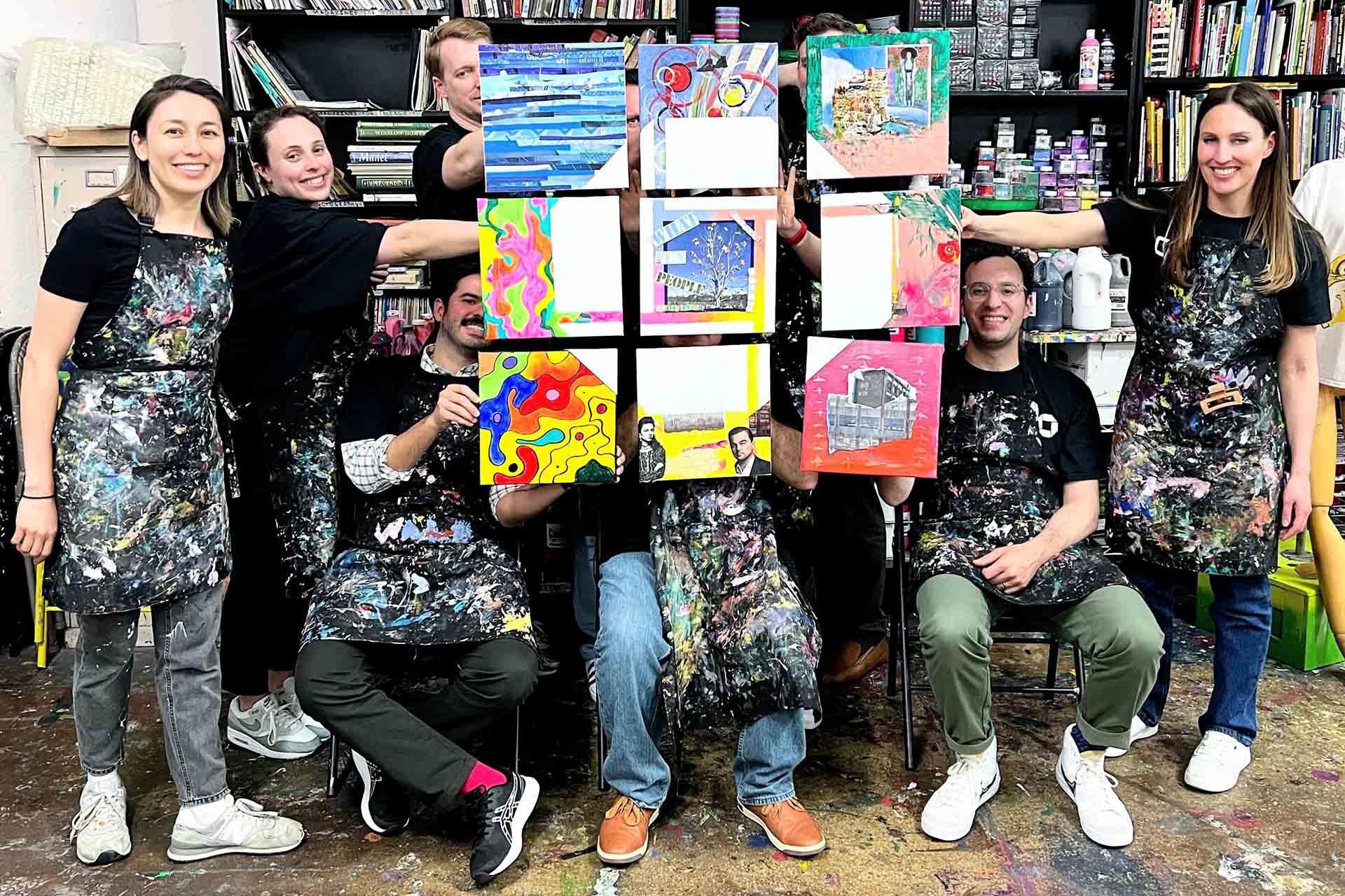 A group of smiling people in paint-splattered aprons hold up colorful geometric paintings in a studio. Shelves with paint bottles are visible in the background. Everyone looks happy and engaged in the art activity.