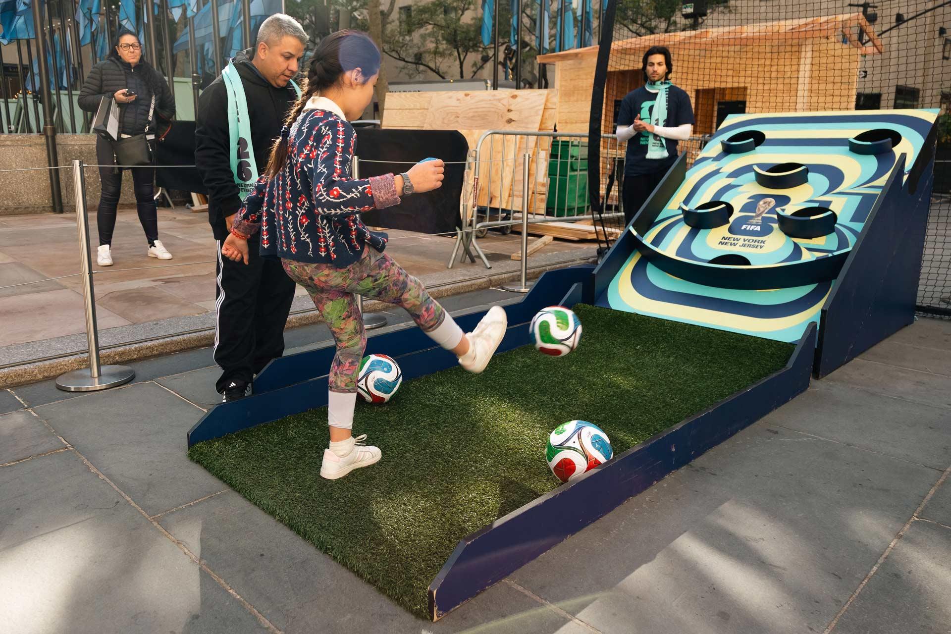 A young girl kicks a soccer ball into a skee-ball style game outdoors, watched by a couple of people. Blue flags and buildings are visible in the background.