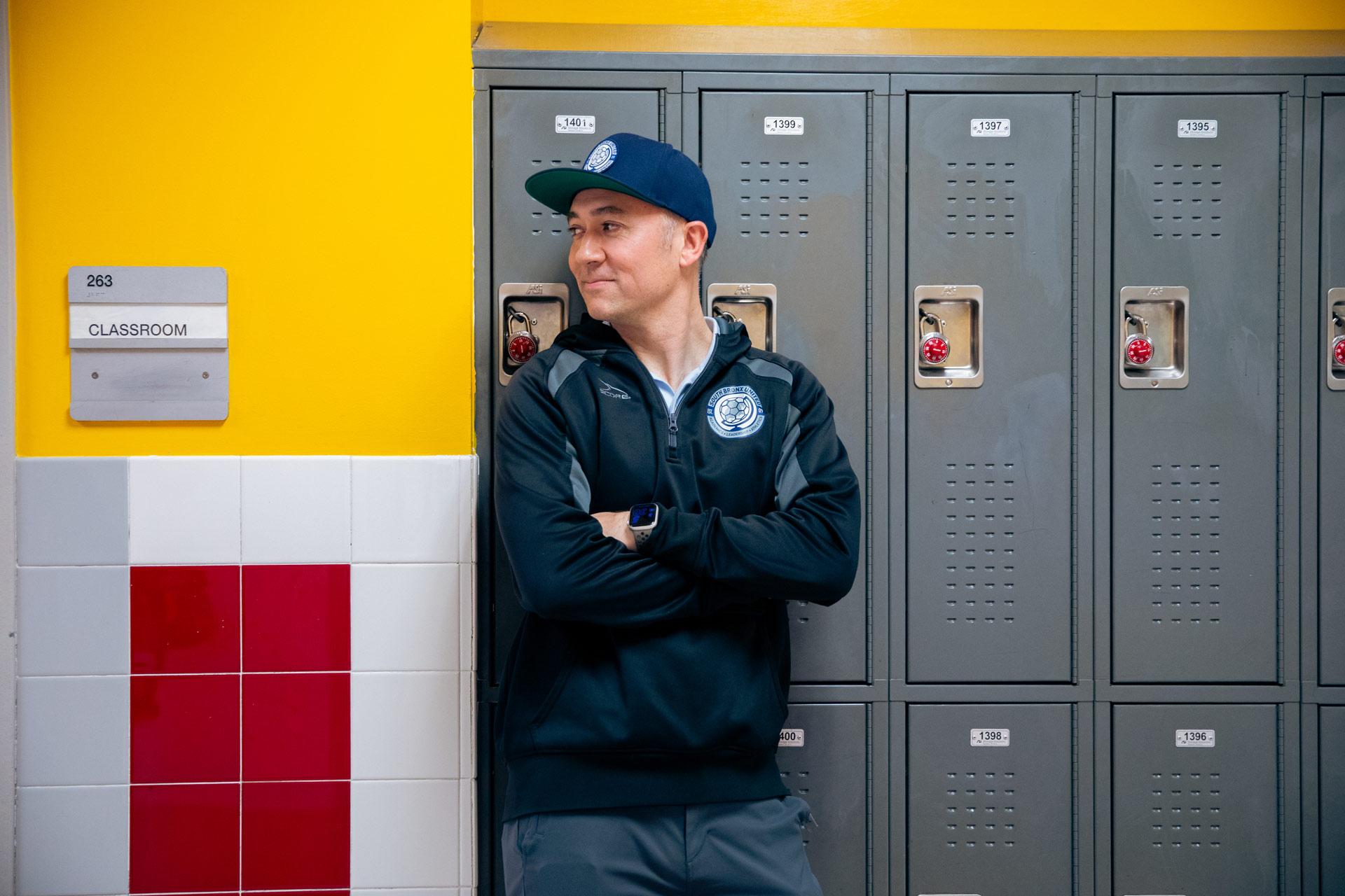 Andrew So in a navy hoodie and cap stands with arms crossed in front of gray lockers, next to a yellow classroom wall with a sign labeled "283 Classroom.
