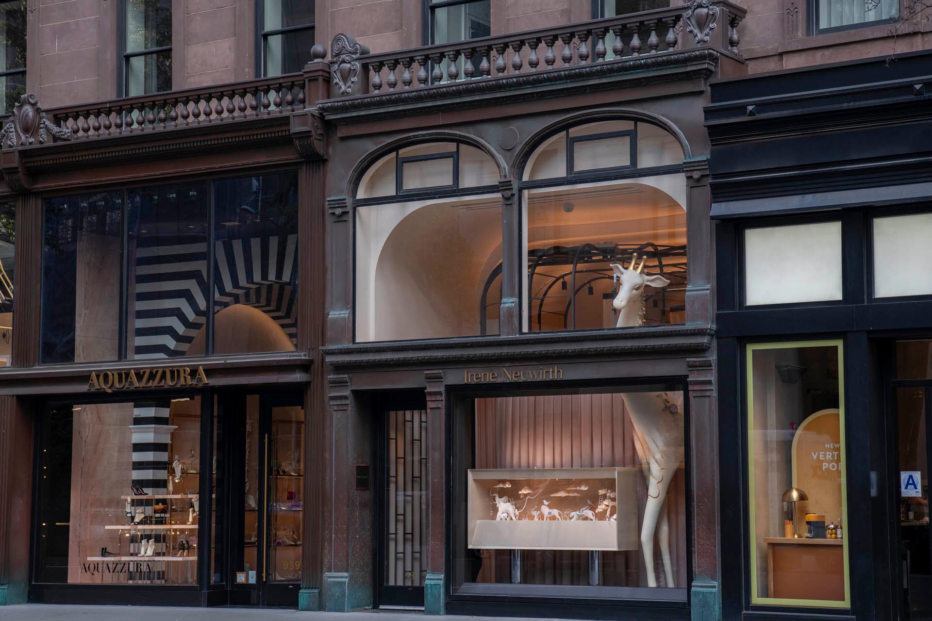 Street view of elegant storefronts with large windows displaying luxury items. A sculpture of a white giraffe is prominently featured in one window. The architecture includes large glass panes and decorative detailing.