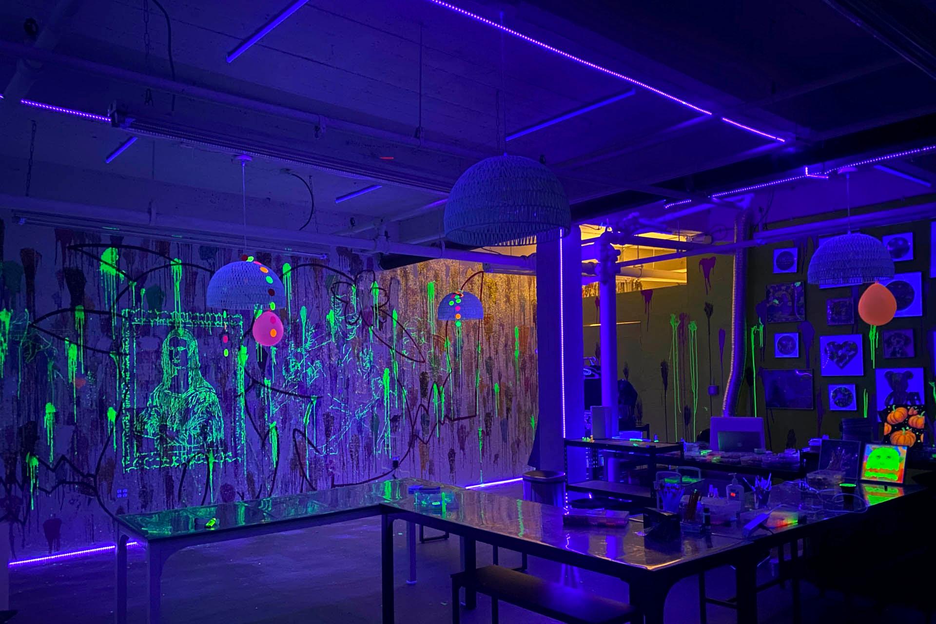 A dark art studio lit by purple and yellow neon lights, with glowing paint splatters on the walls and tables. A Mona Lisa image is drawn in fluorescent paint, with various colorful, glowing decorations hanging from the ceiling.