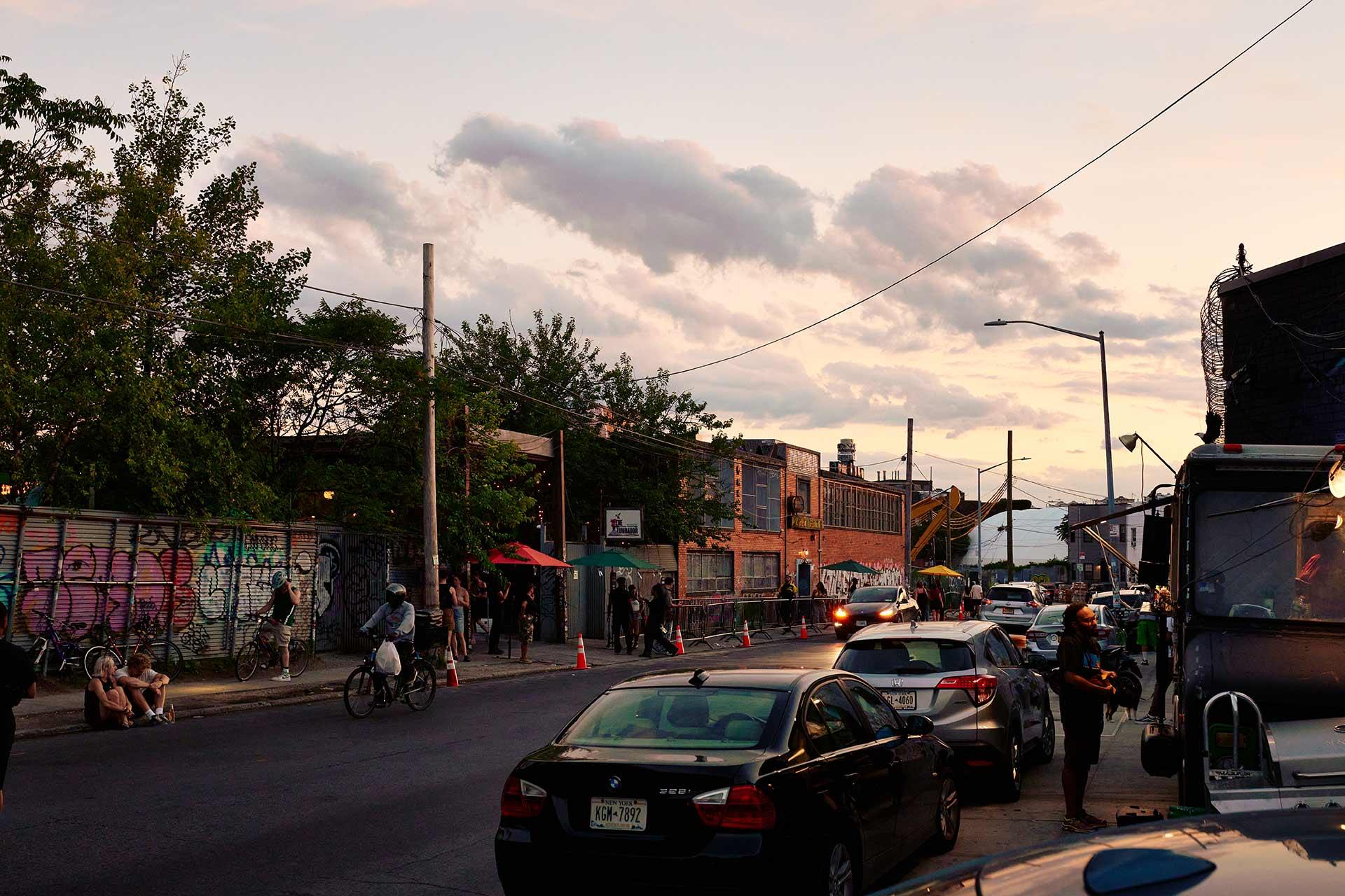 A street scene at dusk with parked cars, a cyclist, and people gathered outside brick industrial buildings with graffiti-covered walls.