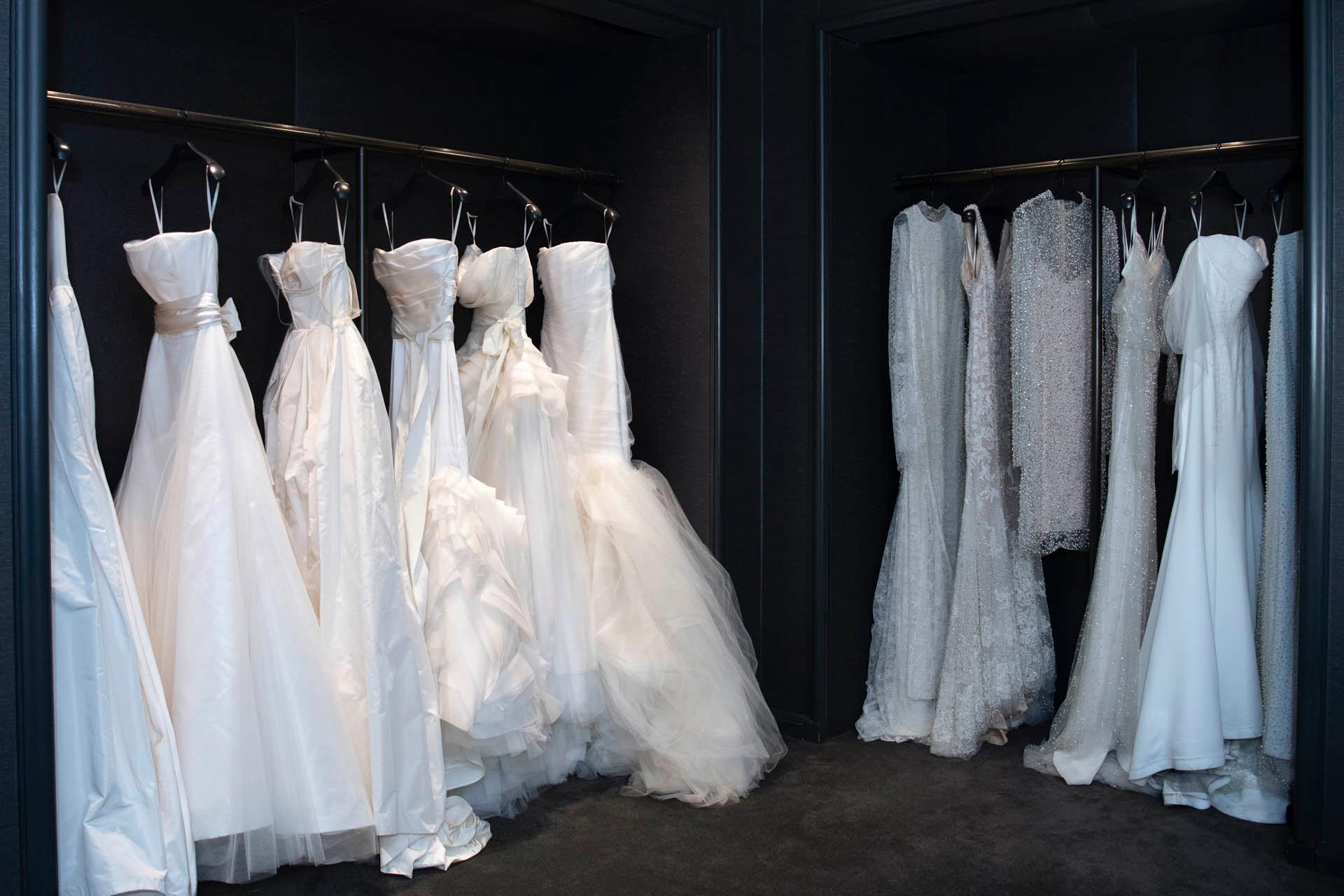 A luxurious bridal shop interior featuring multiple white wedding dresses on display. The gowns, adorned with various designs and textures, hang elegantly on dark racks against a sleek black wall.