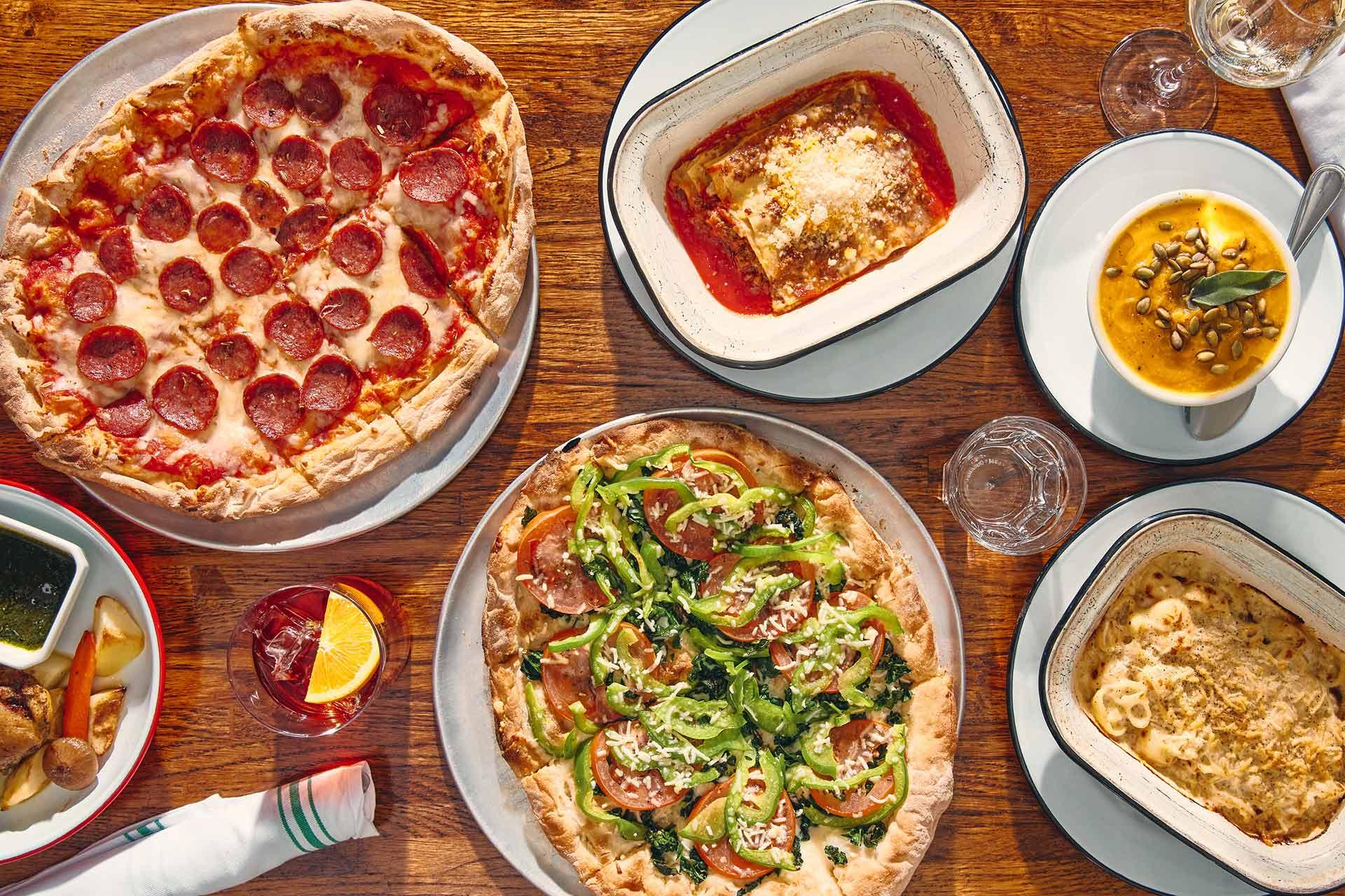 A wooden table set with pepperoni pizza, veggie pizza, lasagna, macaroni and cheese, soup garnished with pumpkin seeds, a cocktail with an orange slice, and a side of bread with dipping sauces.
