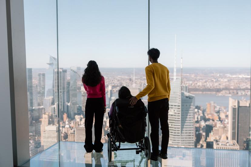 Three people, one in a wheelchair and two standing, look out over a cityscape through large glass windows in a tall building on a clear day.