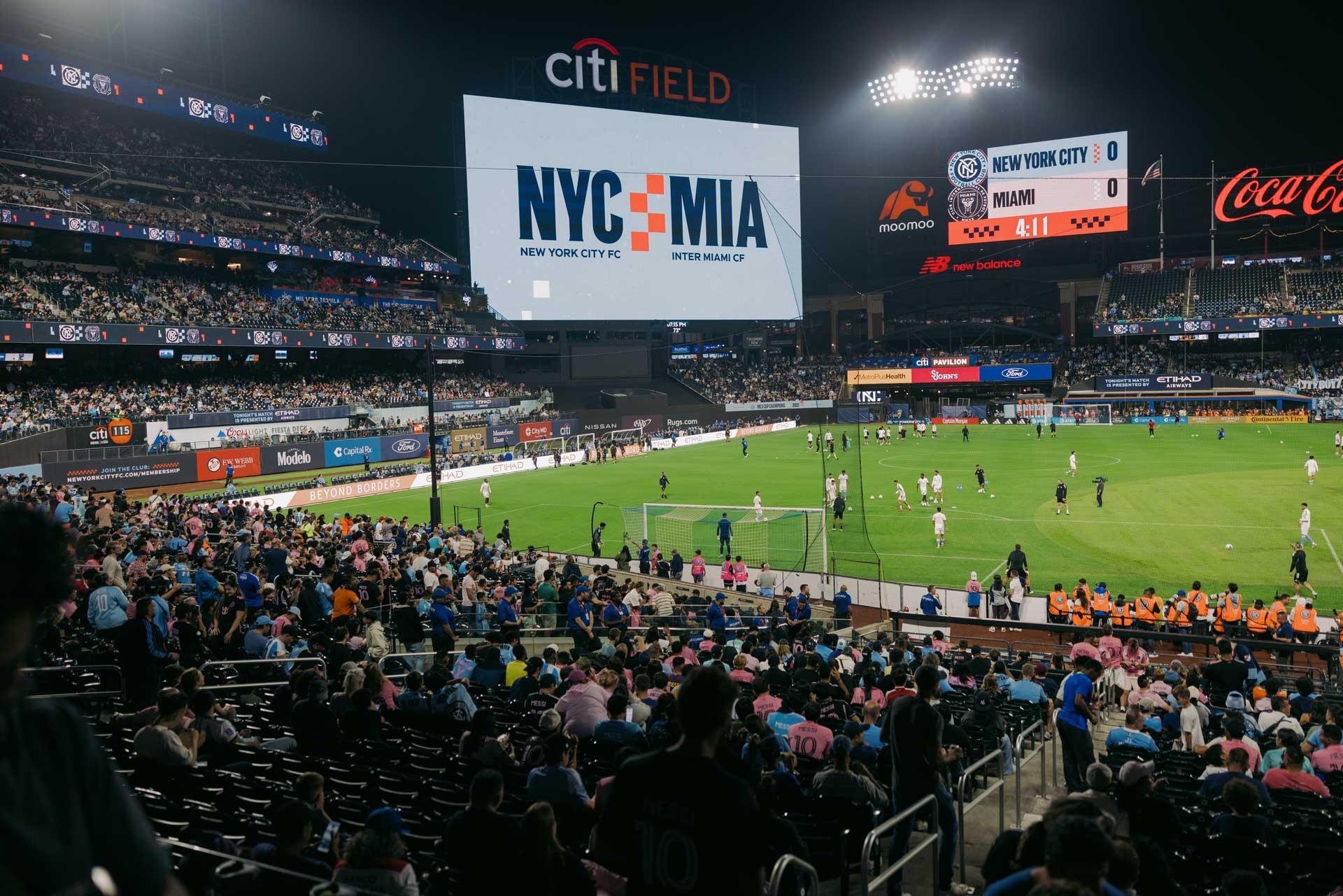 A crowded soccer stadium at night with a large screen displaying "NYC vs MIA". Players are warming up on the field and fans fill the stands at Citi Field for a New York City vs Inter Miami match.