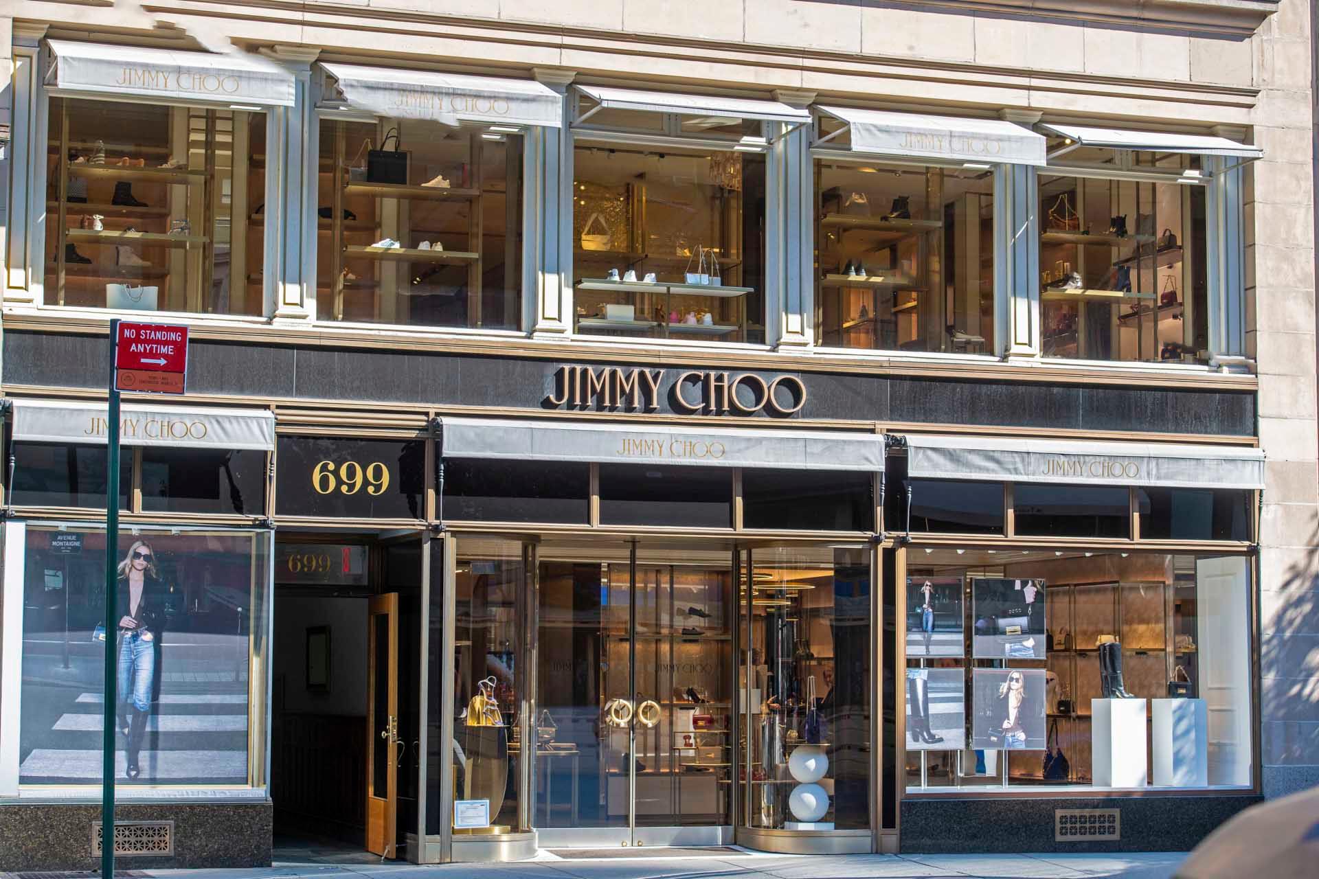 Street view of a Jimmy Choo store. The facade has large glass windows displaying shoes and handbags. The entrance features a single glass door, and the store's logo is prominently displayed above.