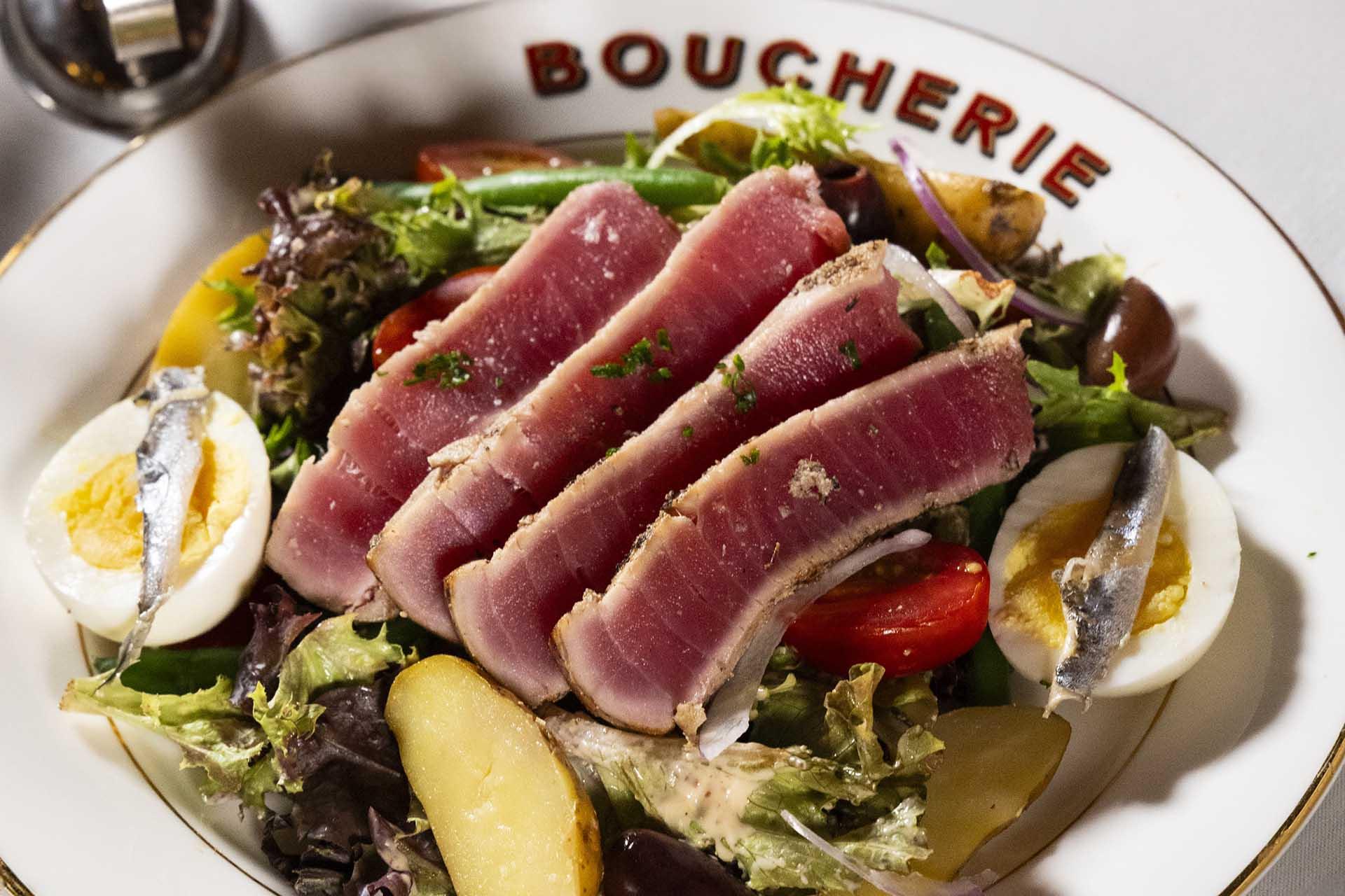 A bowl with seared tuna slices on mixed greens, hard-boiled egg halves, anchovies, tomatoes, potatoes, and green beans, served on a plate labeled “Boucherie.”.