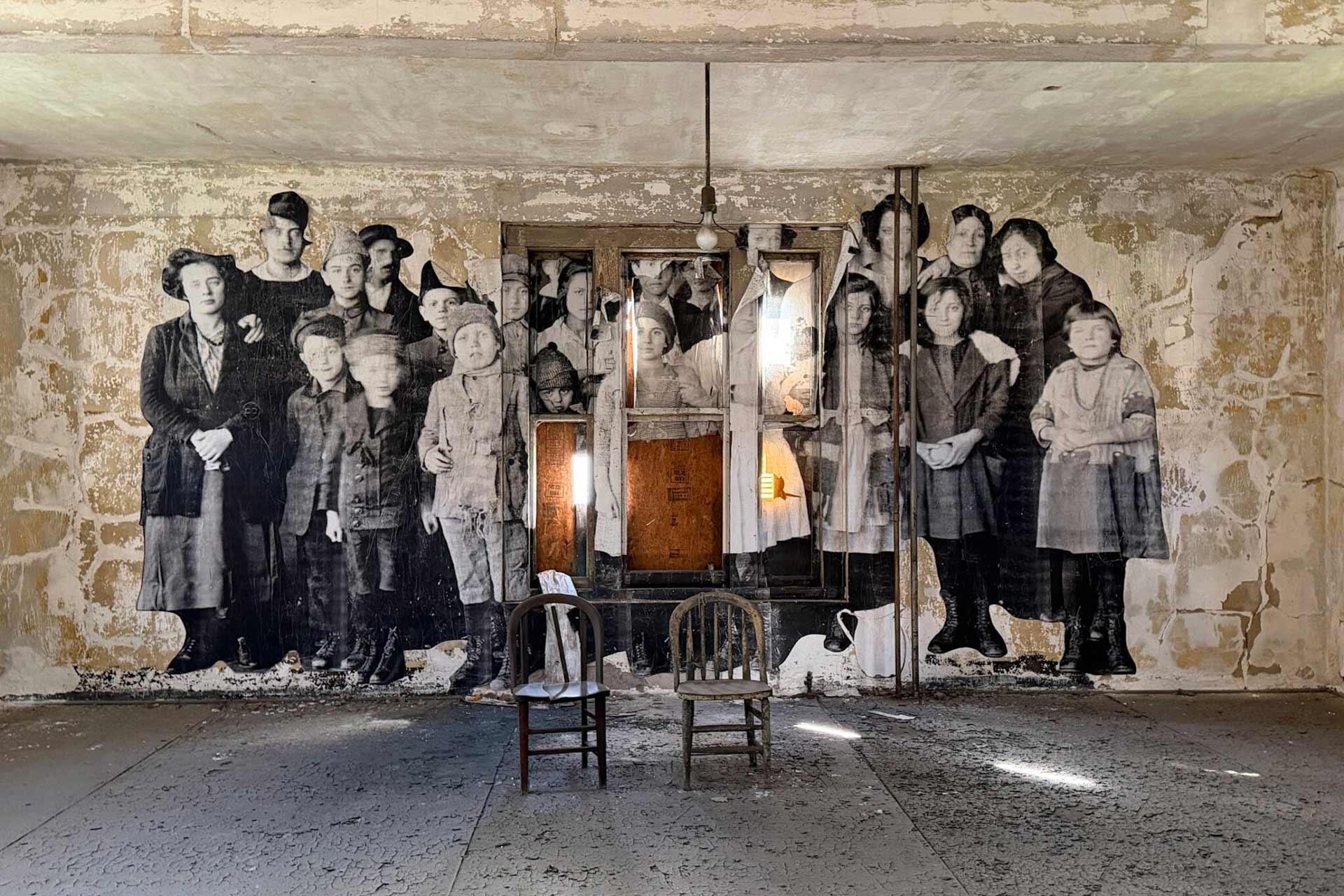 A large, vintage black-and-white photo mural of people, including children and adults, is pasted on a worn, peeling wall in an empty room, with two old wooden chairs placed in front of it.