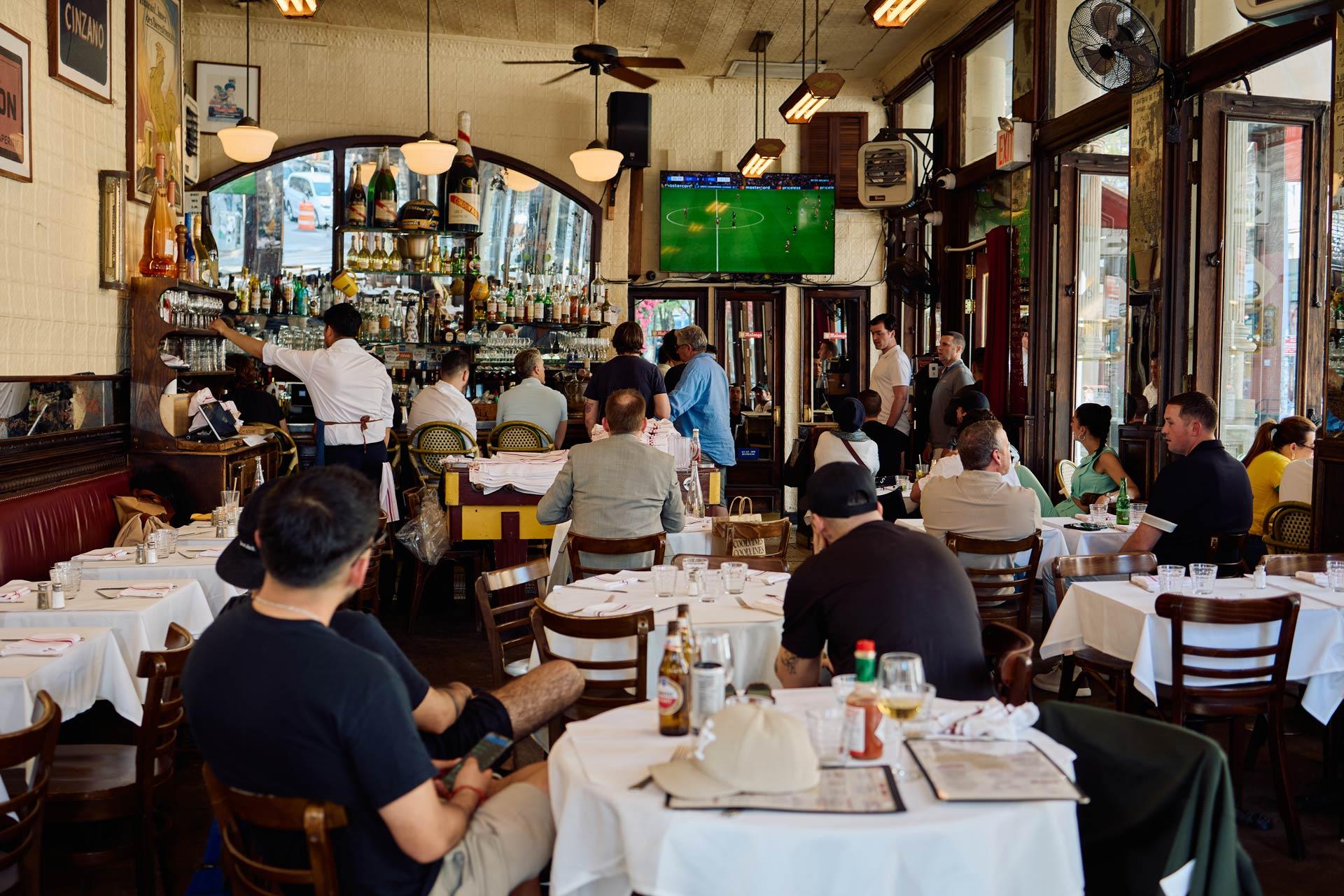 People sit at tables in a busy restaurant, watching a soccer game on a TV mounted above the bar. Some face the screen, while others are eating or talking. The atmosphere is lively and casual, with daylight coming through large windows.