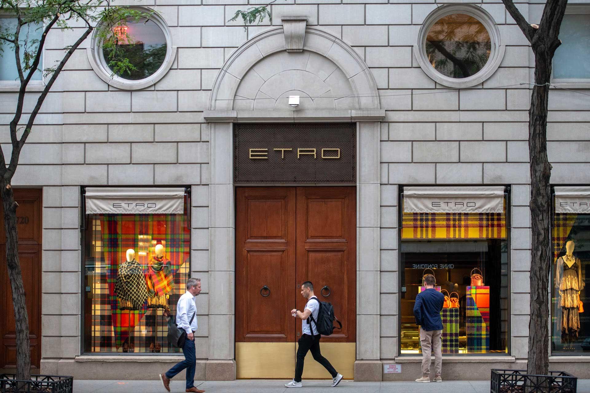 Storefront of Etro on a city street, featuring two display windows with colorful plaid patterns. Three people are walking on the sidewalk. The building has a classic design with large wooden doors and round windows above.