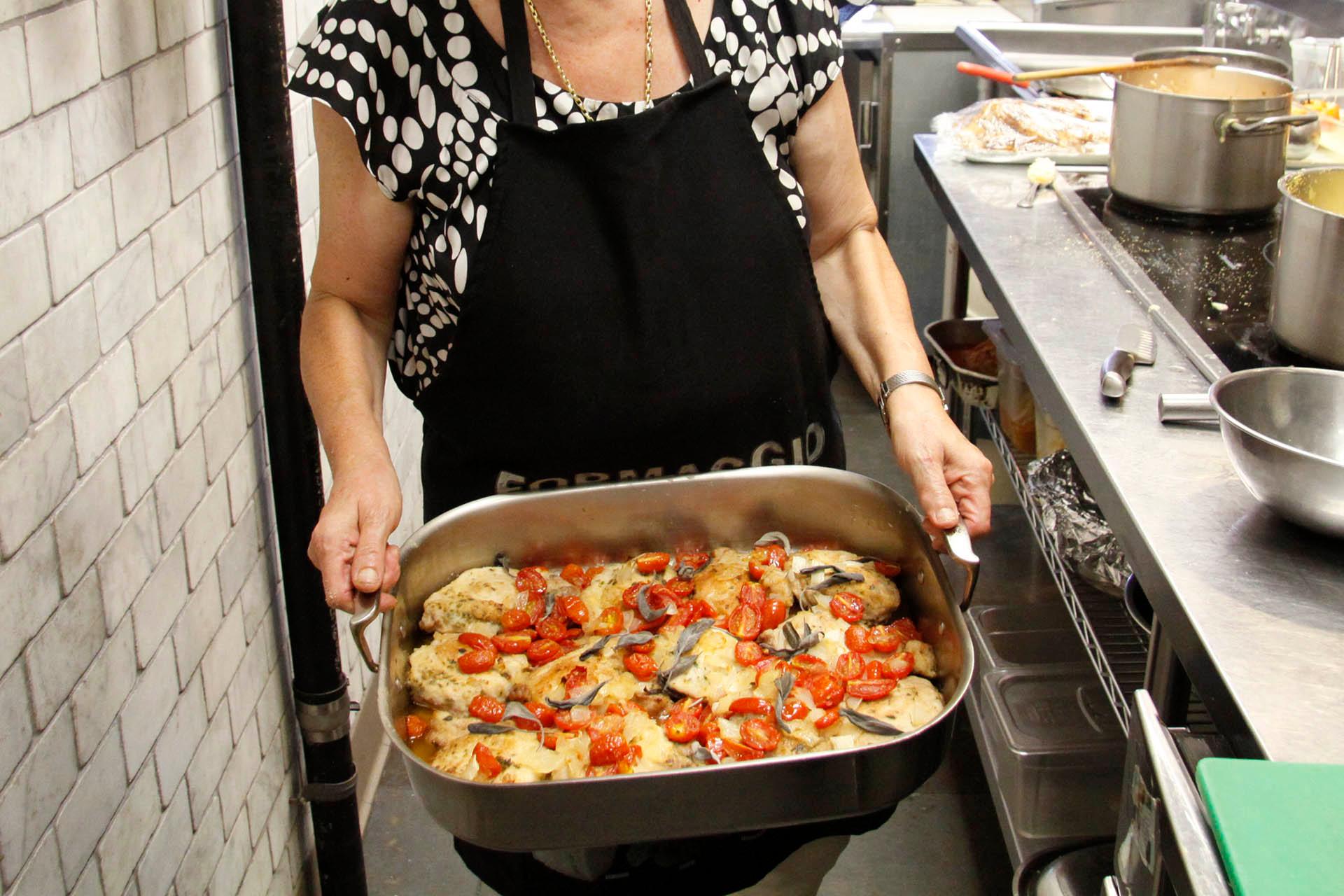 A person wearing a black apron holds a large pan filled with baked chicken, cherry tomatoes, and other vegetables in a kitchen setting. Pots and utensils are visible on the stovetop nearby.