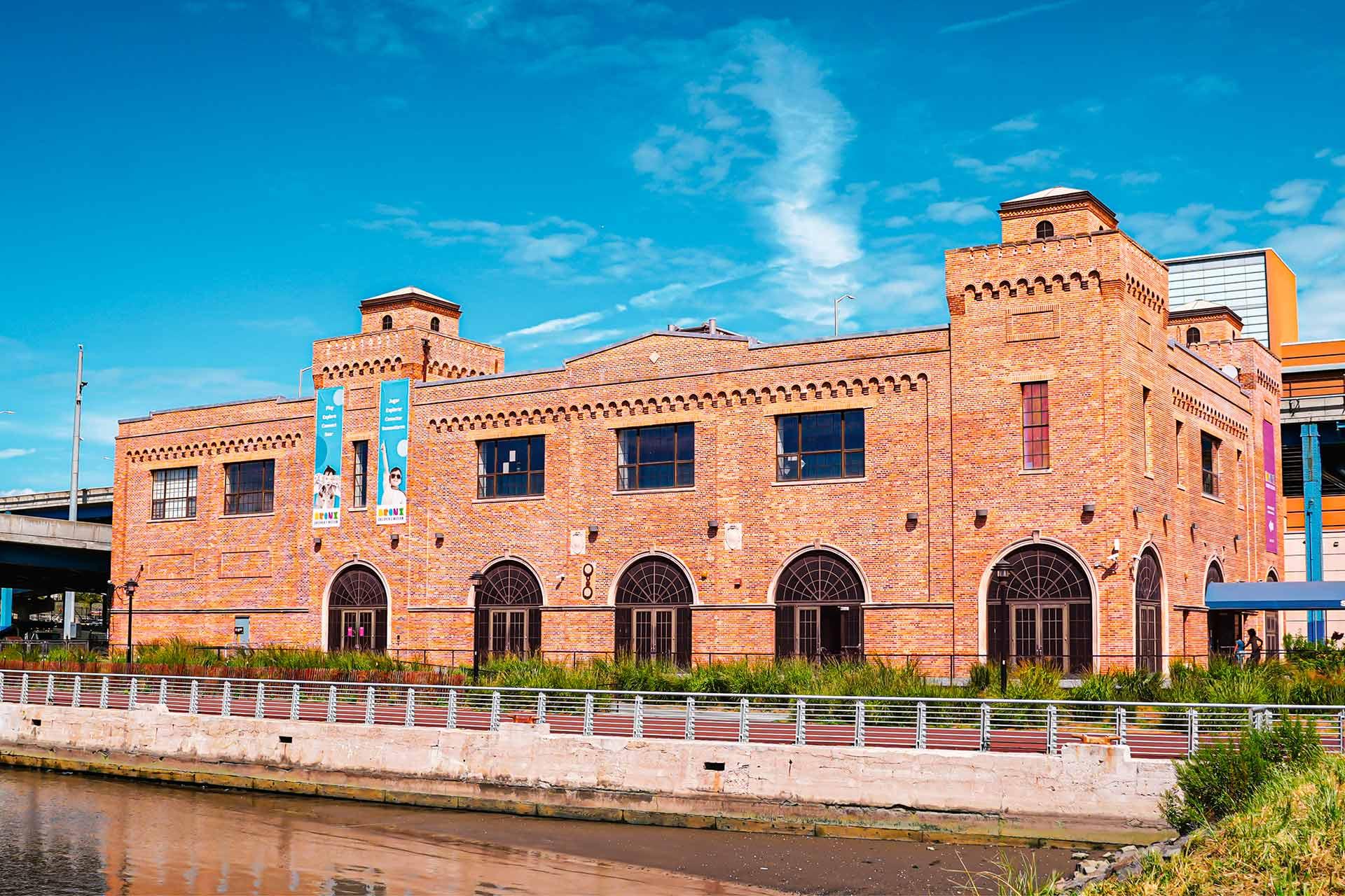 A large brick building with arched windows and castle-like turrets, situated by a waterfront under a bright blue sky. Banners hang on the left side of the building’s facade.