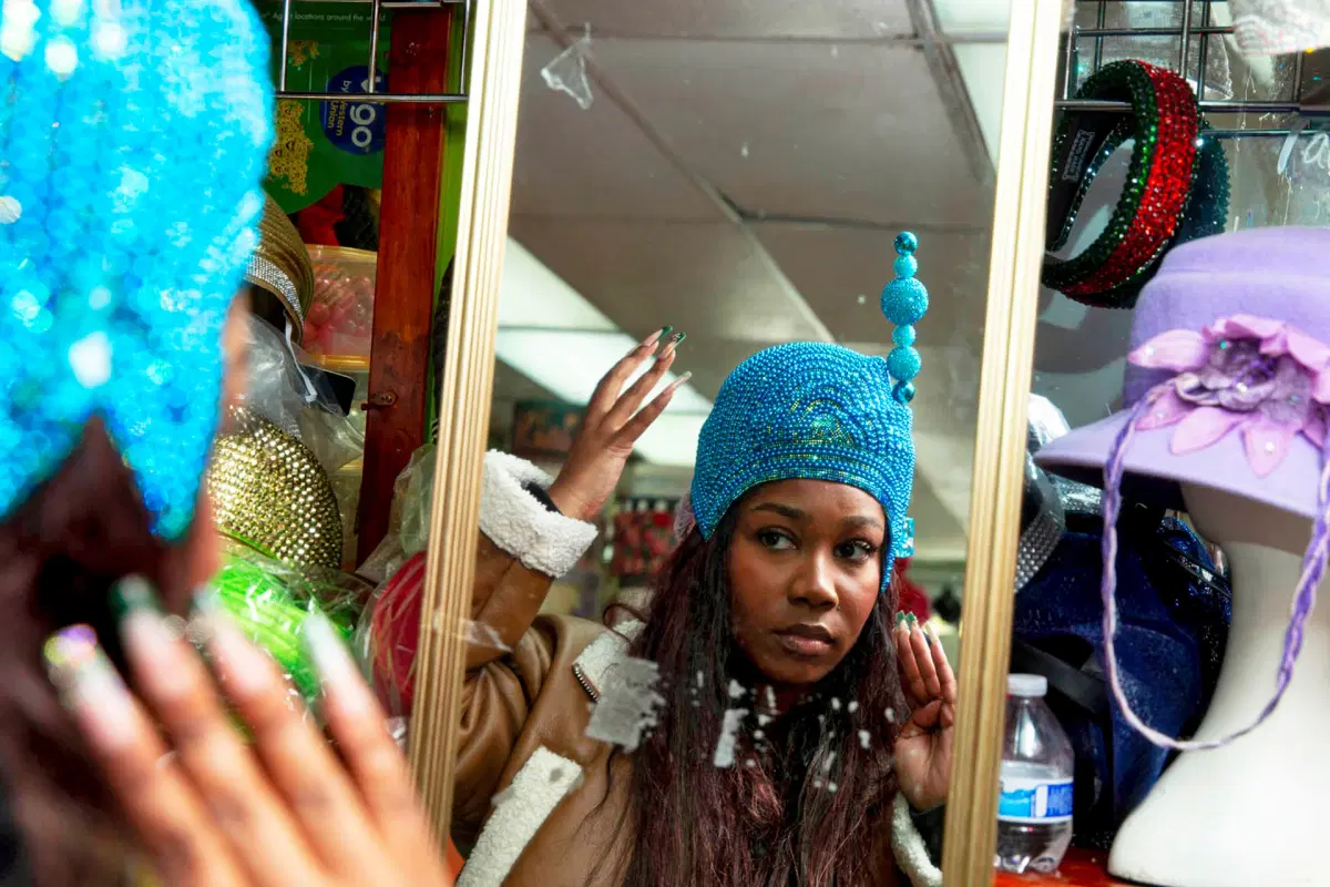 A woman tries on a bright blue beaded hat, looking at her reflection in a store mirror surrounded by colorful hats and accessories.