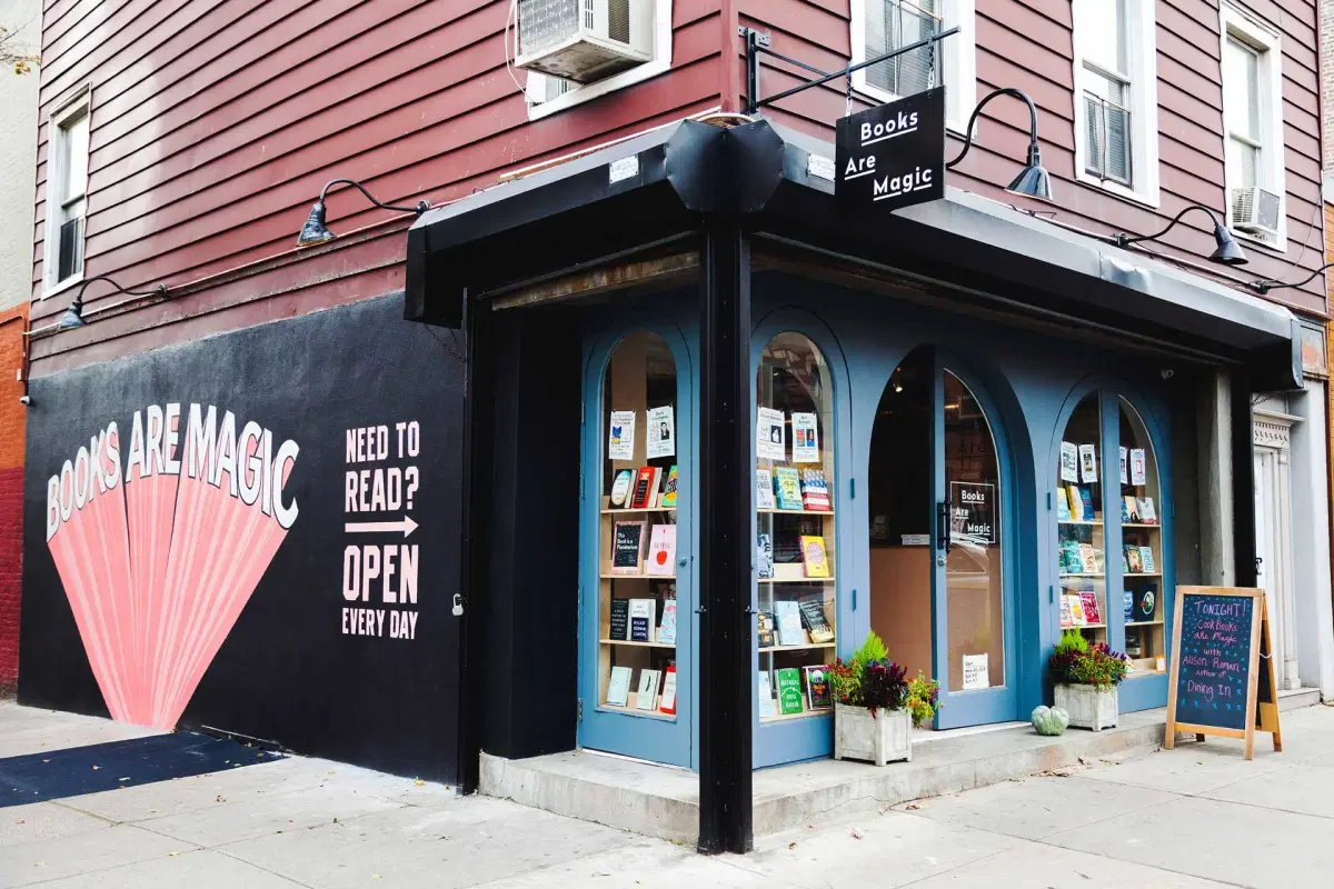 Storefront of a bookstore with curved windows displaying books. The wall reads "Books Are Magic" in large letters and "Need to Read? Open Every Day." The entrance is on a corner with a sandwich board sign outside on the sidewalk.