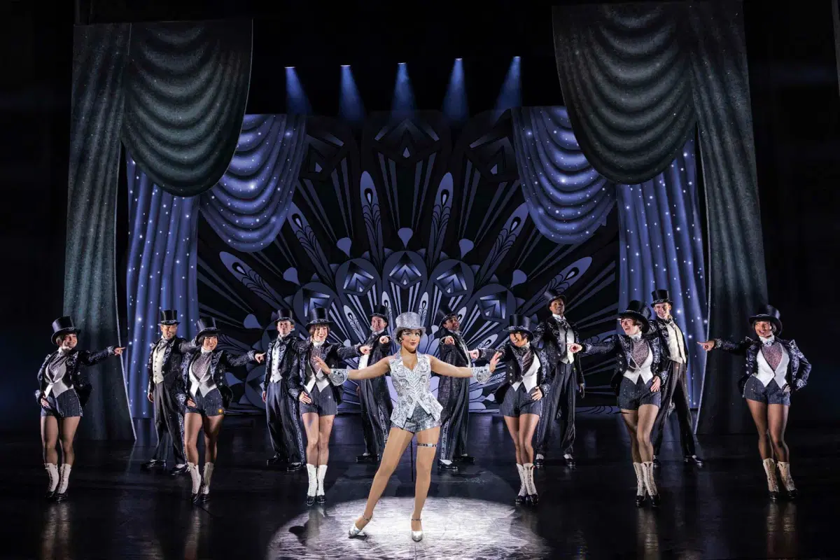 A group of performers is on stage wearing sparkling outfits and top hats. The central performer is highlighted at the front. The backdrop features art deco designs, and the stage is lit with spotlights and draped curtains.