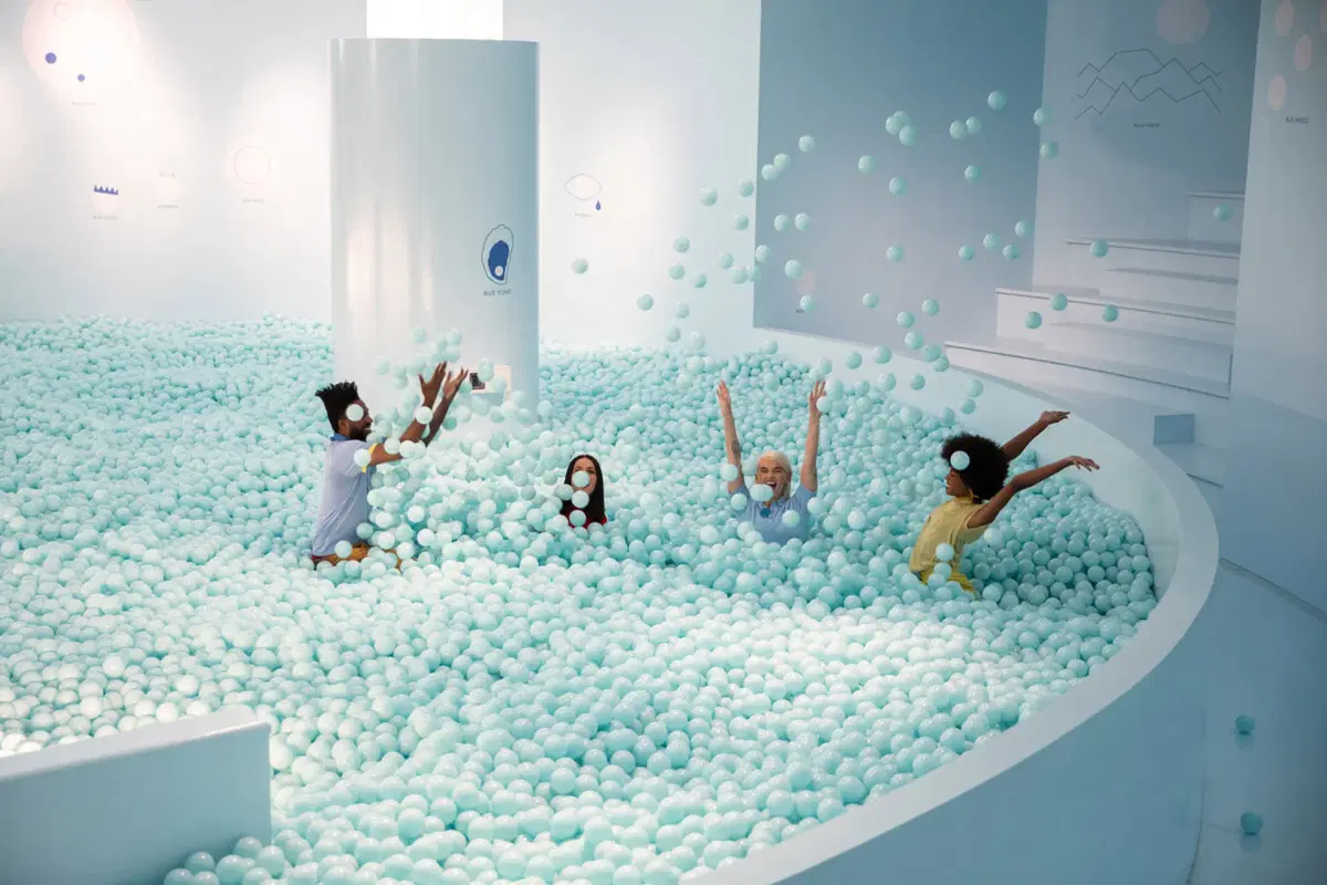 Four children play joyfully in a large indoor ball pit filled with light blue balls, throwing balls into the air. The room is white with minimalist decor and abstract wall art.