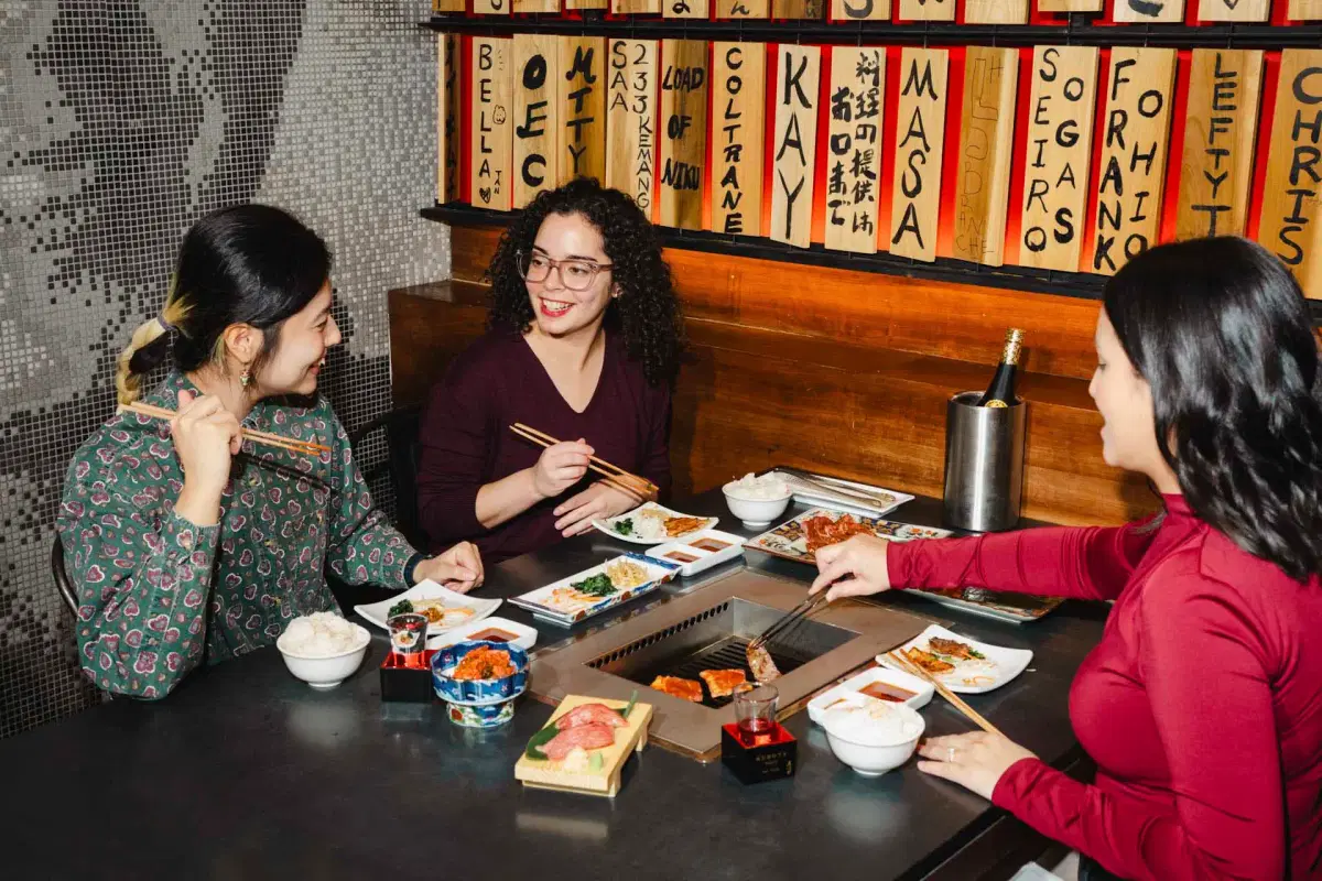 Two women enjoying a meal at a Korean barbecue restaurant. One is using chopsticks and the other is grilling meat. The table is set with various dishes, drinks, and a bottle in an ice bucket. They are seated against a brick wall.