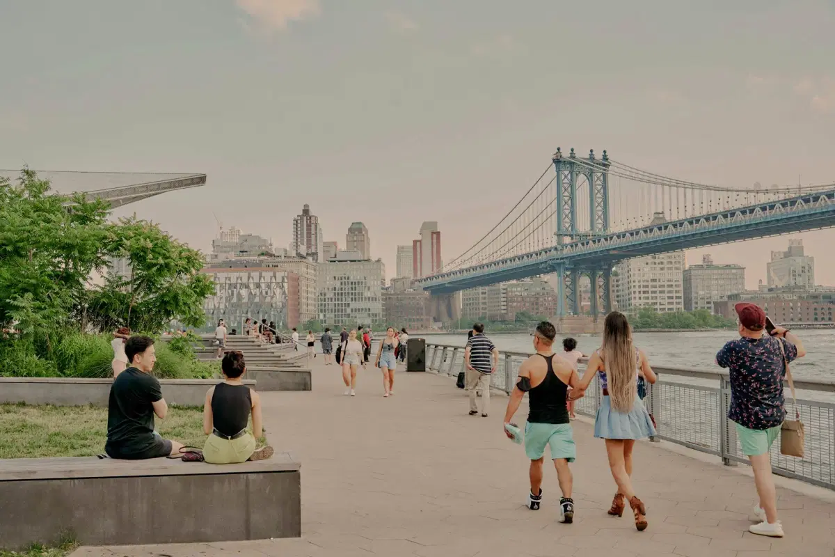 People stroll along a riverside walkway near a large suspension bridge. Some sit on benches, while others walk. Tall buildings are visible in the background under a cloudy sky.
