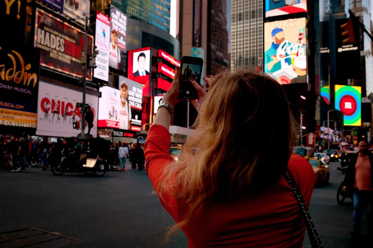 A person with long hair and an orange top takes a smartphone photo in Times Square, which is filled with bright billboards and advertisements.
