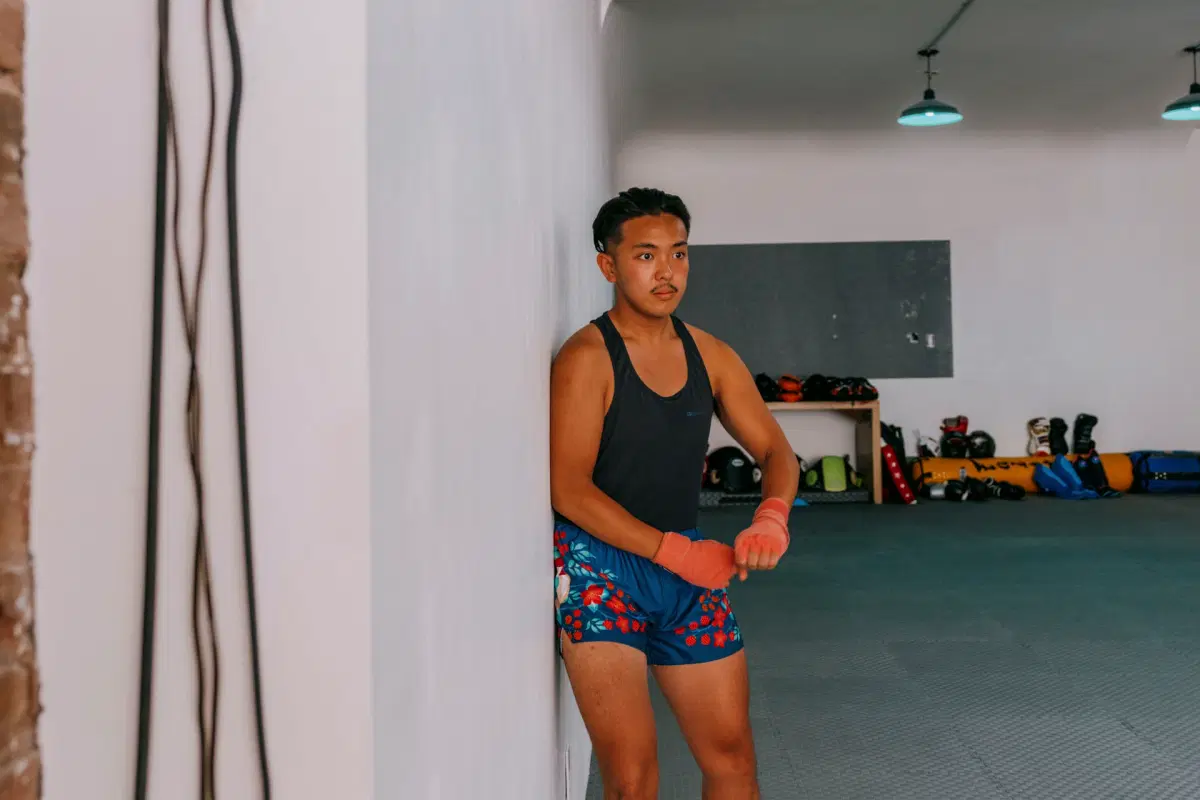 A person with long hair is leaning against a white wall in a gym, wearing a dark tank top and patterned shorts. Their hands are wrapped, and they appear focused. Gym equipment is visible in the background.