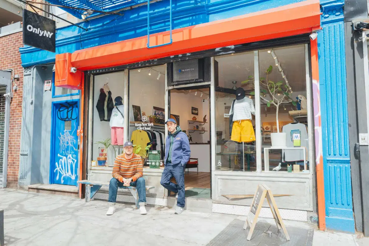 Julian and Micah wearing casual clothes stand in front of the OnlyNY storefront with a glass display window, mannequins, and street art graffiti on the adjacent wall.