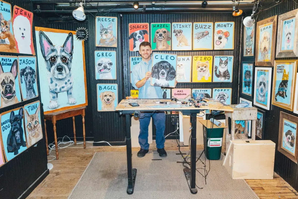Ben Lenovitz stands in his studio holding a dog portrait. The walls behind him are covered with colorful, framed dog paintings, each labeled with a different name. Art supplies are on a desk in front of him.