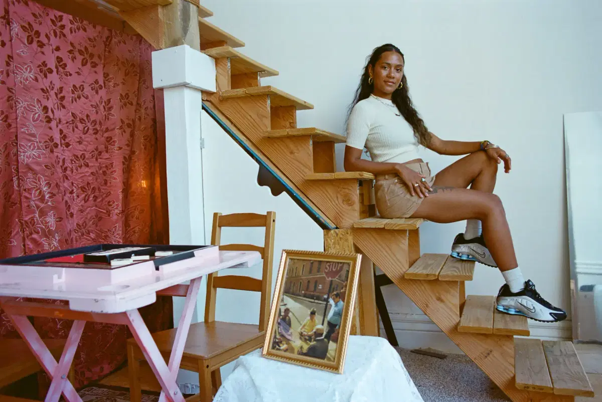 Djali Brown-Cepeda with long hair sits on wooden stairs indoors, wearing a white top, beige shorts, and sneakers. Nearby are a wooden chair, a pink table, and a framed photo on a small white-covered table. A mirror rests against the wall.