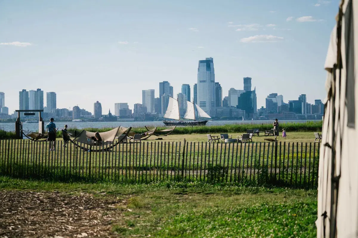 A grassy park with hammocks and people relaxing, bordered by a wooden fence. A sailboat is on a nearby body of water, with a city skyline featuring tall buildings in the background under a clear sky.