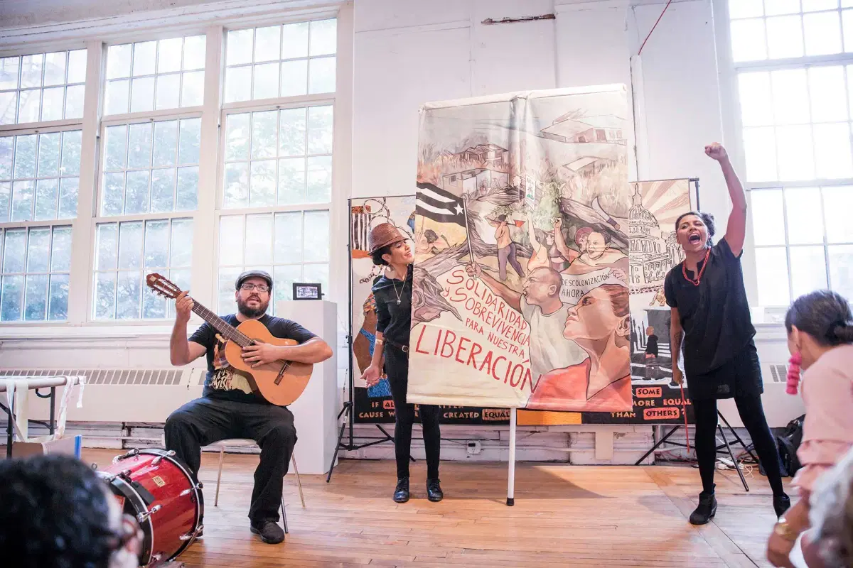 Three performers stand in front of a colorful banner about liberation inside a bright room; one plays guitar, one holds the banner, and one raises a fist while speaking passionately to an audience.