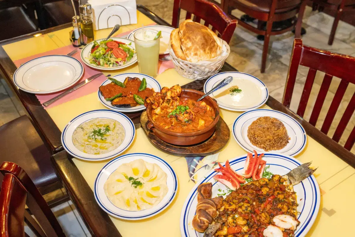 A table set with various Middle Eastern dishes, including grilled fish, stews, hummus, salads, sauces, fried appetizers, flatbreads in a basket, and a glass of lemonade. The table is surrounded by wooden chairs.