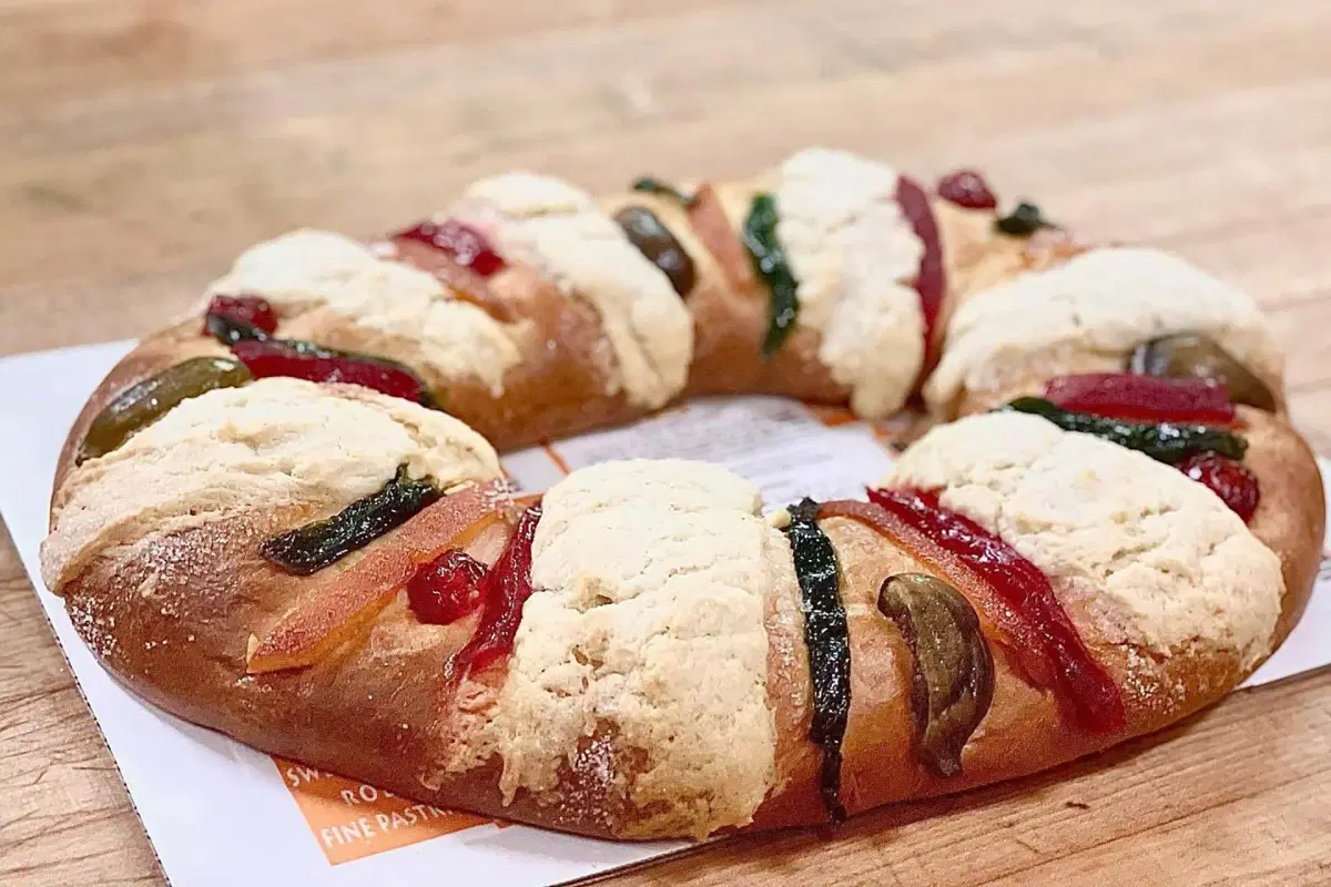 A round, ring-shaped sweet bread decorated with colorful candied fruits and sugar topping, placed on a wooden surface.