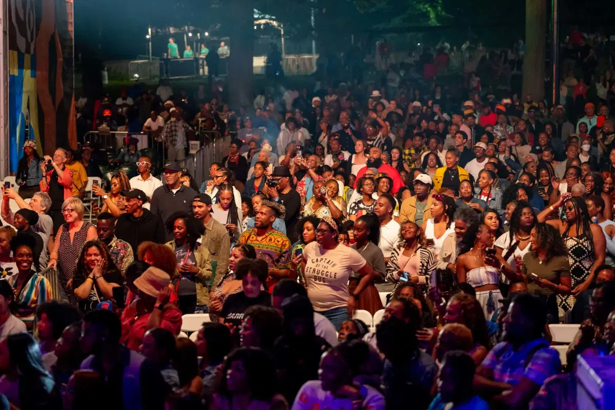 A large, diverse crowd of people stands and sits outdoors at night, enjoying a lively event. Many are smiling, clapping, or raising their hands, with trees and lights visible in the background.