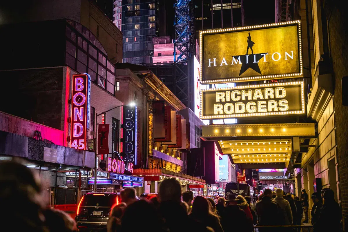 A crowded nighttime street in New York City’s theater district, featuring the brightly lit Richard Rodgers Theatre marquee with “Hamilton,” and neon signs for Bond 45 on nearby buildings.