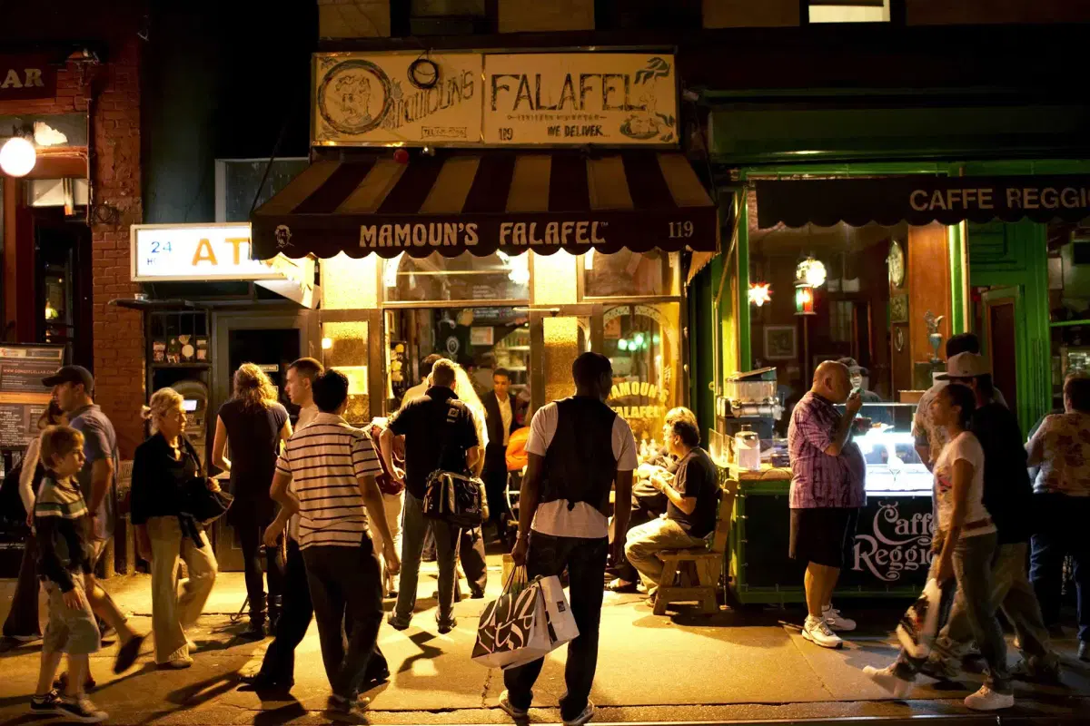 A busy nighttime street scene shows people gathered outside Mamoun’s Falafel and nearby Caffe Reggio. The restaurants are lit warmly, and people are eating, chatting, and walking along the sidewalk.