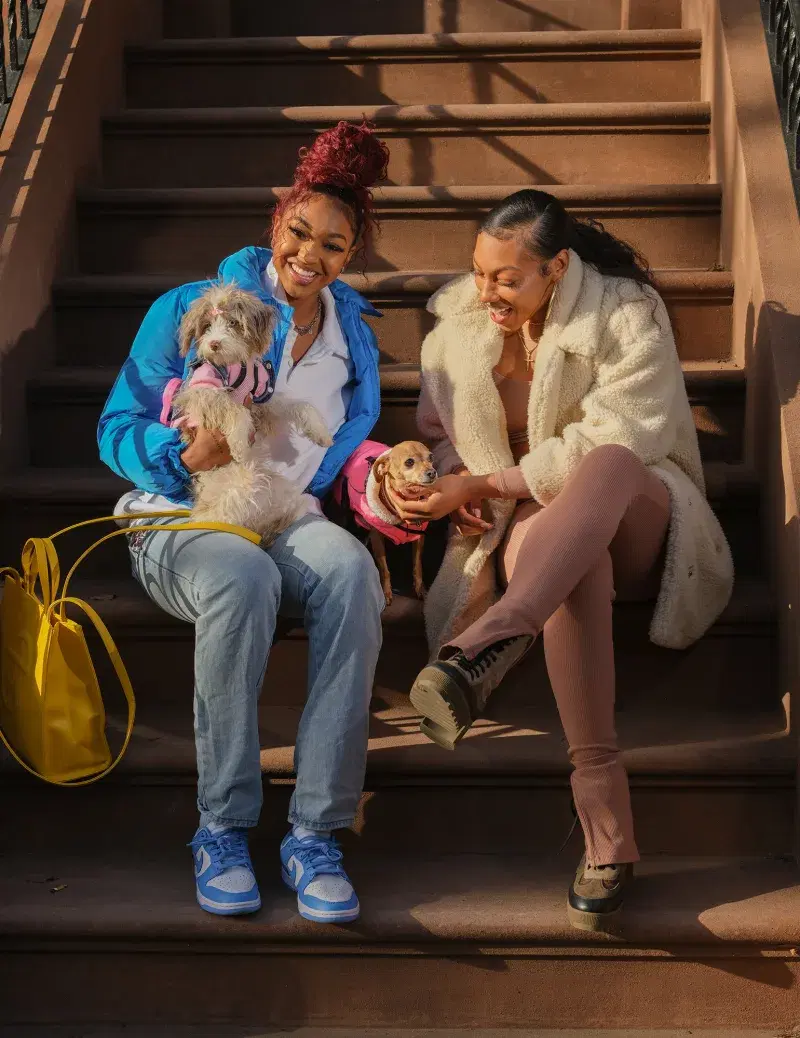 Portrait of Betnijah Laney and Didi Richards of the New York Liberty, sitting on a stoop with two dogs, in New Brooklyn